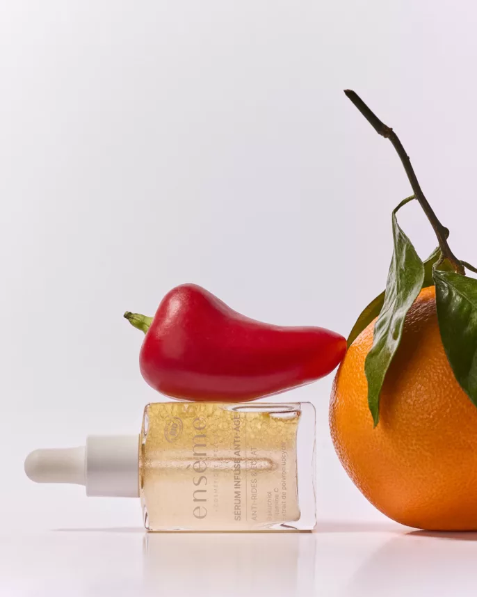 Photographie en studio d'un flacon de sérum anti-âge Ensème allongé, entouré d’un poivron rouge et d’une orange avec feuilles, sur fond blanc minimaliste.