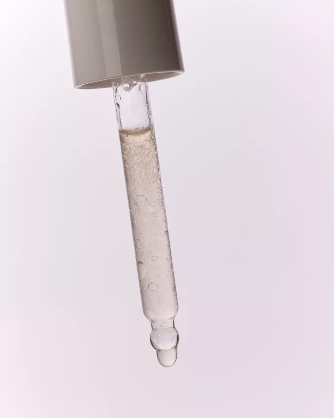 Goutte de sérum cosmétique suspendue au bout d’une pipette transparente sur fond rose pâle.