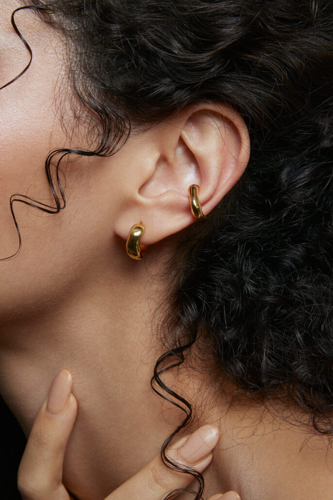 Zoom sur une oreille ornée de deux boucles d’oreilles dorées, photographie de bijoux en studio à Lyon — mise en valeur des textures, boucles naturelles et manucure nude.