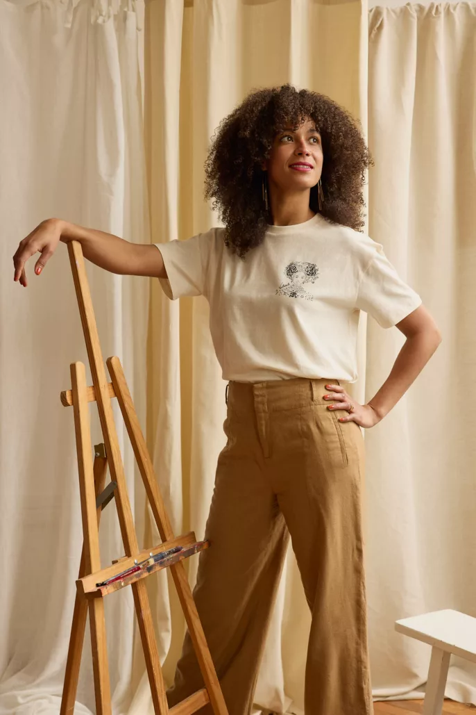 Femme souriante posant en studio à côté d’un chevalet, vêtue d’un t-shirt blanc imprimé et d’un pantalon beige – photo de mode réalisée à Lyon.
