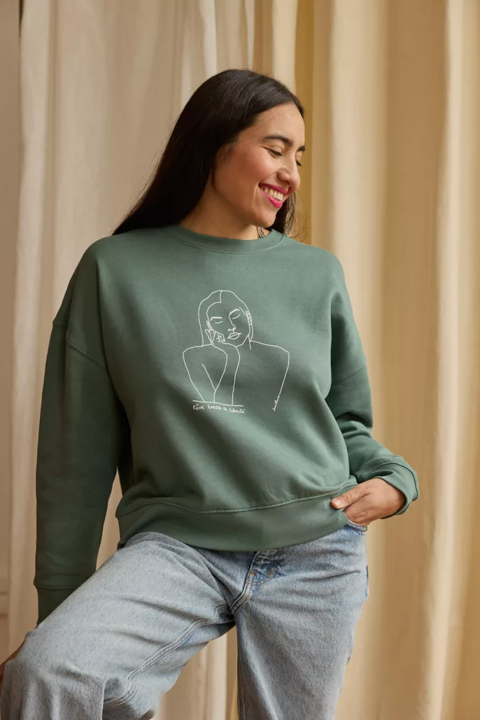 Portrait d’une femme souriante en studio, portant un sweat vert illustré d’un dessin minimaliste – photographie mode à Lyon pour marque engagée.