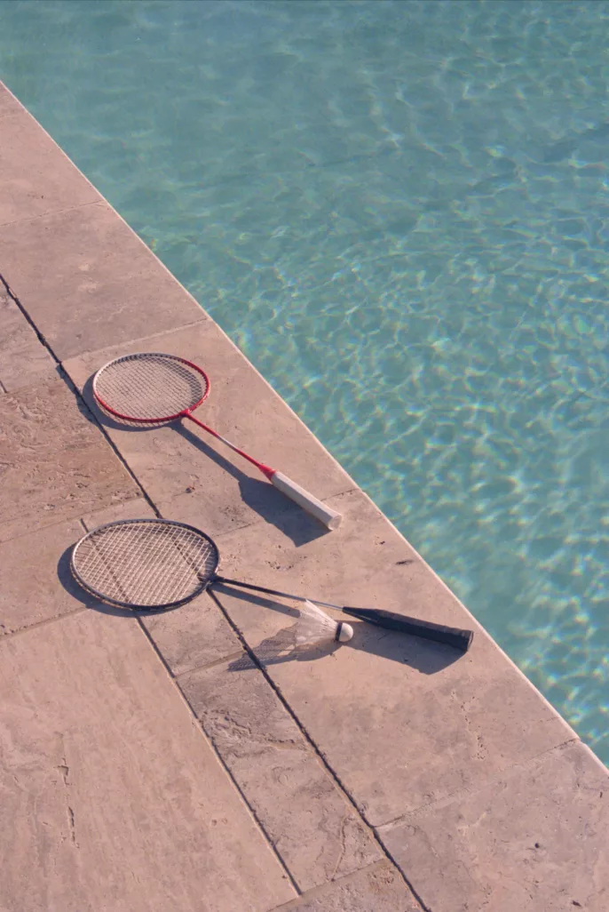 Deux raquettes de badminton et un volant posés au bord d’une piscine, lumière naturelle d’été.