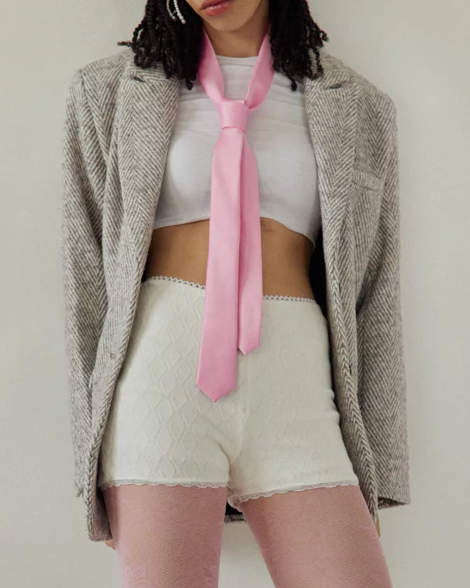 Portrait mode en intérieur d’une femme portant une cravate rose, un blazer à chevrons, un crop top blanc et un short en maille avec collants roses.