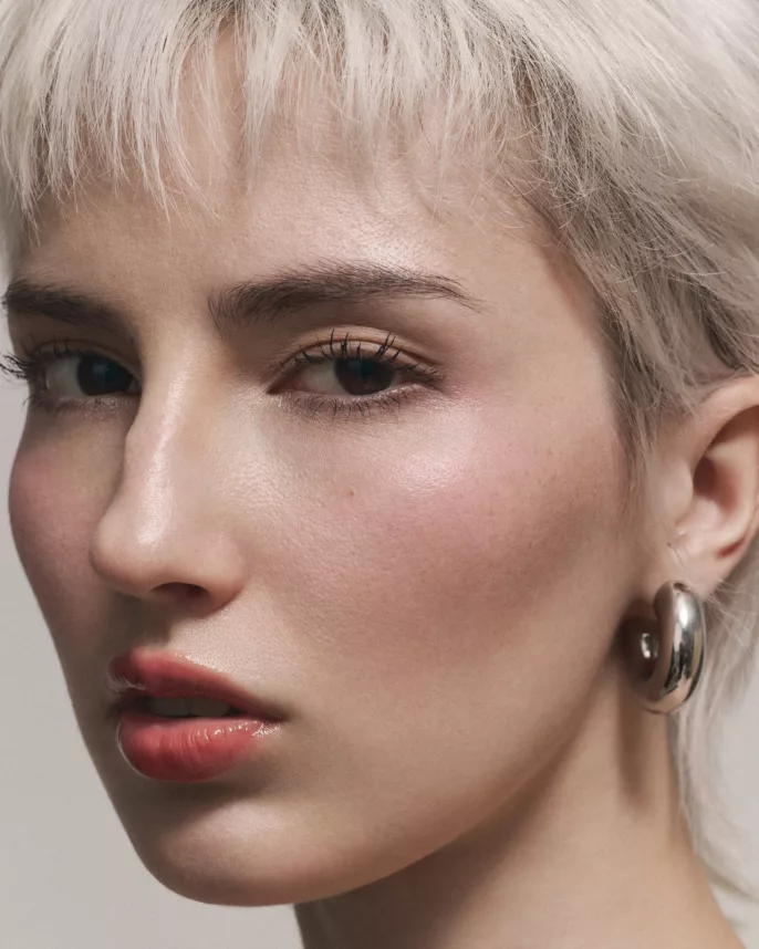 Portrait serré d’une femme aux cheveux courts platine, maquillage nude et boucle d’oreille épaisse métallique.