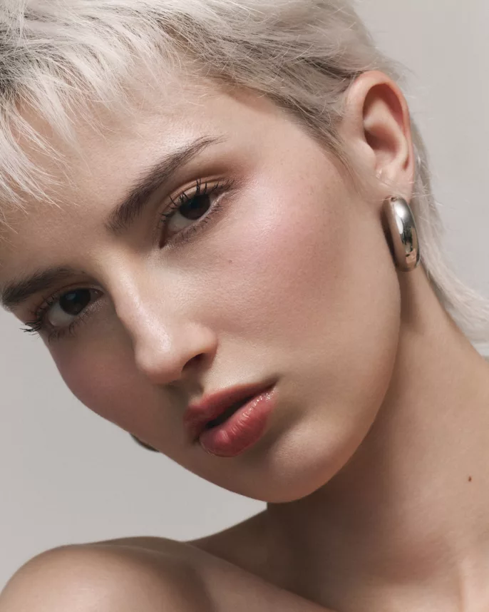Portrait beauté d’une femme aux cheveux courts platine, maquillage nude et boucle d’oreille métallique.