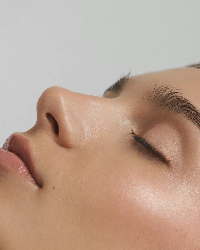 Close-up d’un visage féminin allongé, avec peau lumineuse et yeux fermés.