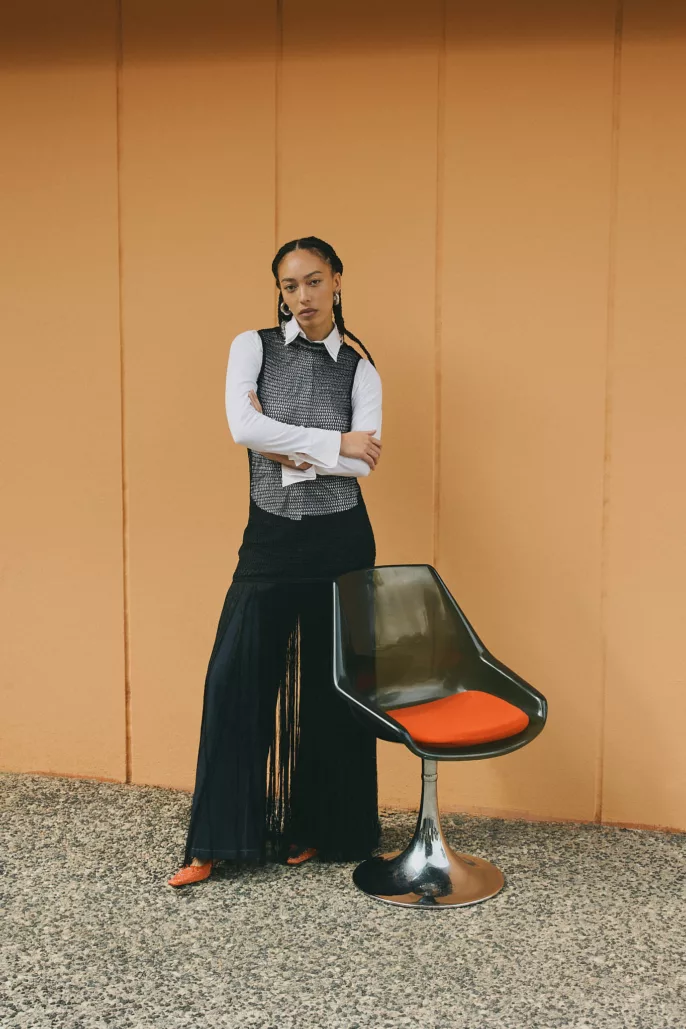 Femme debout en tenue noire face à un mur orange, aux côtés d’une chaise design – Photographe mode éditorial Lyon