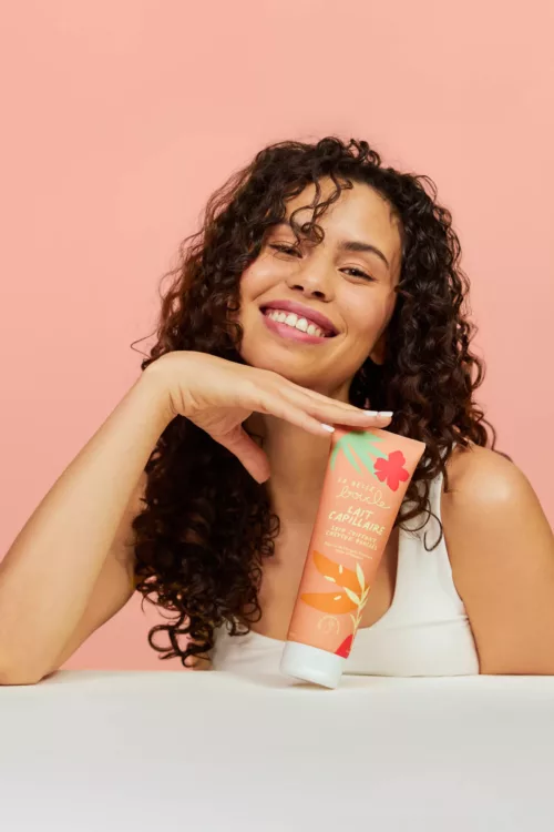 Modèle aux cheveux bouclés souriante avec le lait capillaire La Belle Boucle posé devant elle — photographe beauté Lyon, Stéphanie Lorang
