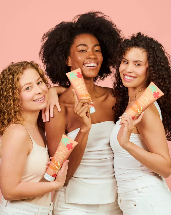 Trois femmes aux cheveux bouclés tenant le lait capillaire La Belle Boucle sur fond rose — photographe beauté Lyon, Stéphanie Lorang