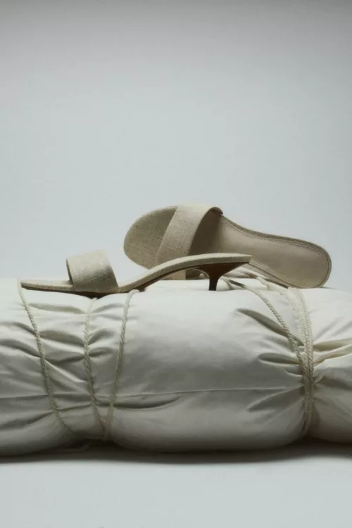 Mules en lin crème posées sur oreiller ficelé sur fond gris clair — photographie éditoriale studio Lyon, Stéphanie Lorang